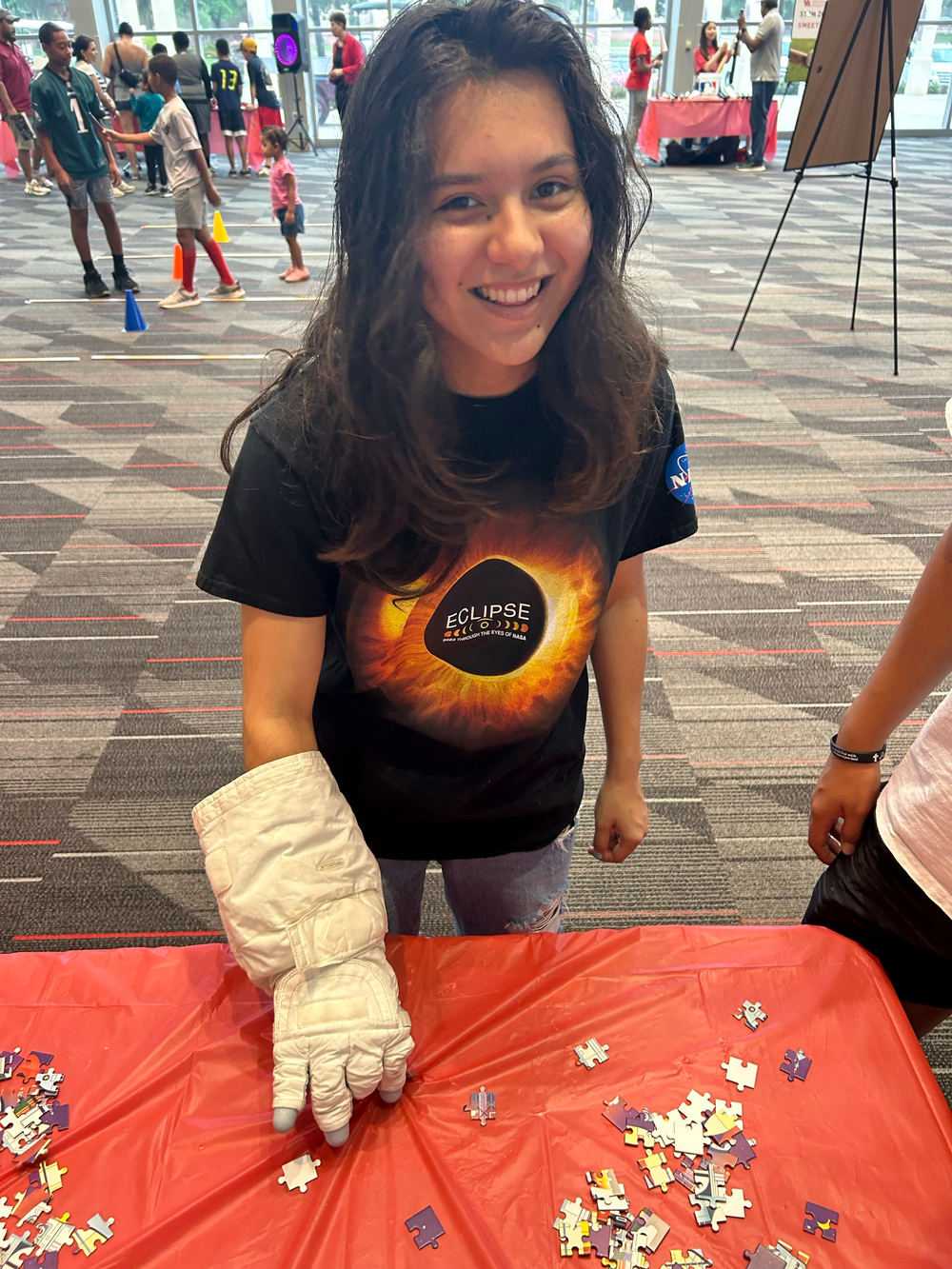 A young girl wearing a space glove and a black shirt with jeans smiles while putting puzzle pieces together. 